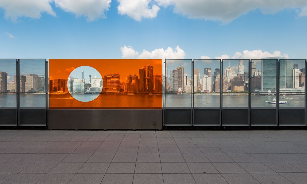 An orange artwork panel installed in a glass railing on the East River waterfront, framing the Manhattan skyline behind it.